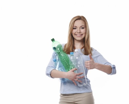 A young woman with long, straight, blonde hair and a cheerful expression is holding multiple transparent plastic bottles, some with green or blue caps, against her chest. She is wearing a light blue, long-sleeved blouse with rolled-up sleeves and beige trousers. The bottles appear to be empty and are of varying sizes, including a large green one, a clear one with a blue cap, and smaller bottles. She is standing on a plain white background, with her left arm giving a thumbs-up gesture, indicating approval. The scene is well-lit, and the focus is on her positive expression and the collection of plastic bottles she is holding, which relates to environmental awareness and waste management themes often associated with rubbish removal services. The image conveys a message of environmentally conscious waste disposal and recycling efforts in a professional and neutral manner, suitable for inclusion on a website such as commercialwastesutton.co.uk under the 'rubbish' service category, serving the Sutton area.
