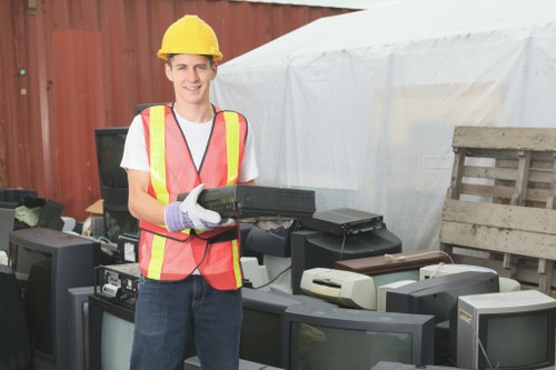 A young male waste management worker wearing a yellow safety helmet, a bright yellow and red high-visibility vest over a white T-shirt, and work gloves, is standing outdoors on a paved area. He is smiling and holding a black, rectangular electronic device or component. In front of him, there are multiple discarded electronic appliances, including old televisions with large screens, visible in various shades of grey, black, and cream with some showing signs of wear. The background features a red wooden fence and a white tent-like structure. The surrounding environment suggests a disposal or recycling area for electronic waste, consistent with rubbish removal services such as those offered by Commercial Waste Sutton in the Sutton area, potentially near the postcode SM suburb. The scene is well-lit, indicating daytime, with a focus on waste collection or clearance of electronic and general rubbish materials.