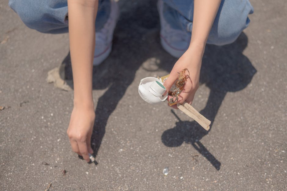 A person kneeling on a paved outdoor surface, holding a small white ceramic bowl with a rounded shape and glossy finish, partially filled with tangled natural fiber and debris. The person's left hand 